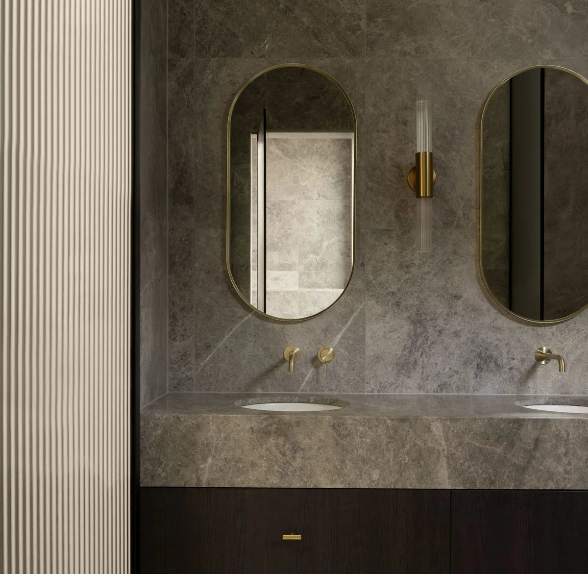 Modern bathroom vanity with a dark wood double sink cabinet, gray stone countertop and wall, two oval mirrors, and gold-tone faucets and hardware.